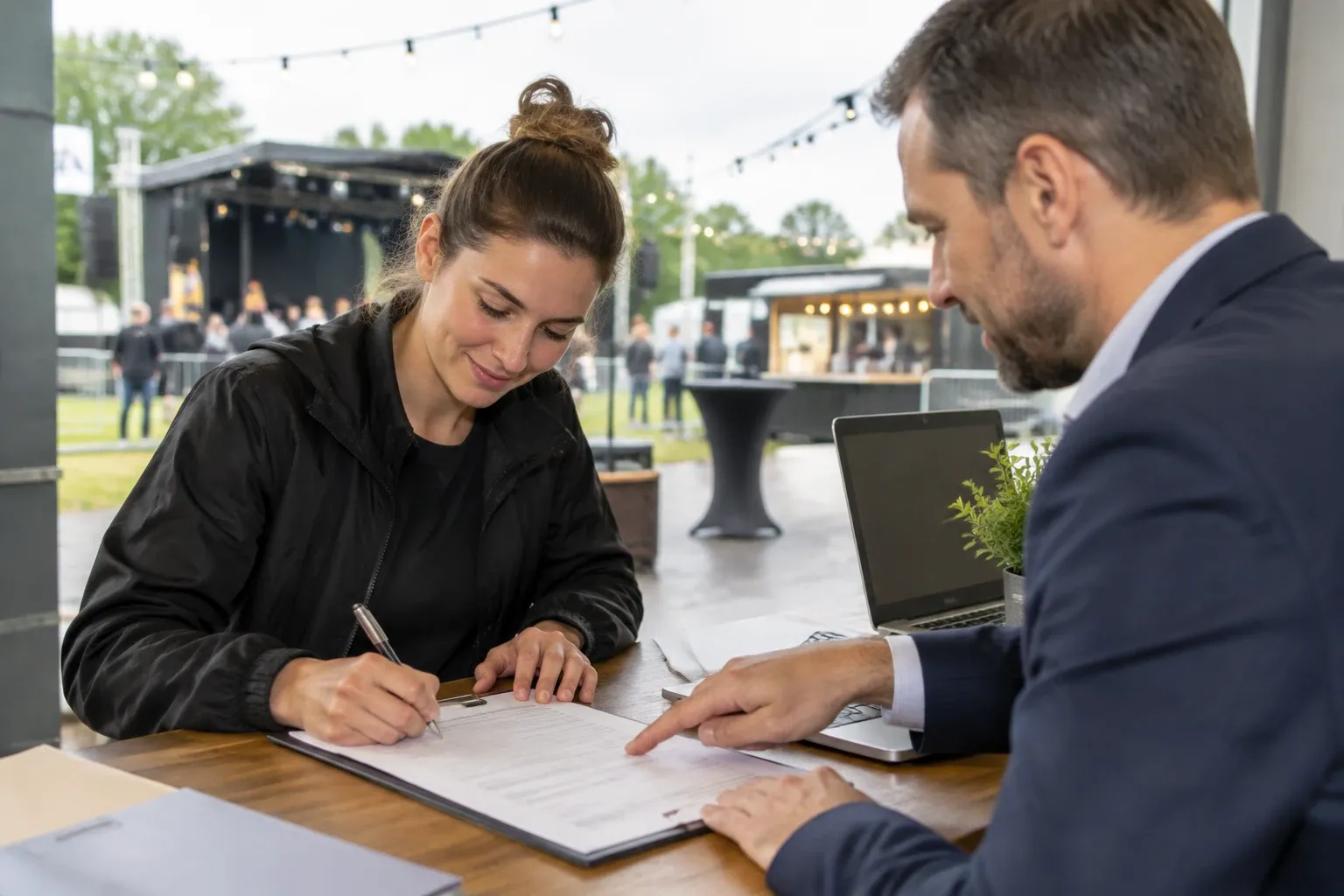 Vrouw tekent vergunningformulier aan tafel buiten, man wijst op document, podium op achtergrond