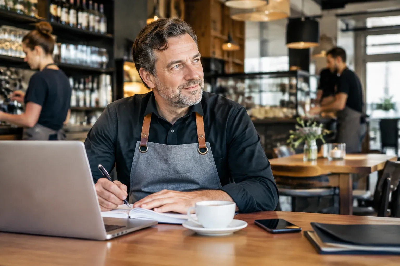 Koffiekop en laptop op tafel, nadenkende barista in schort schrijft in notitieblok